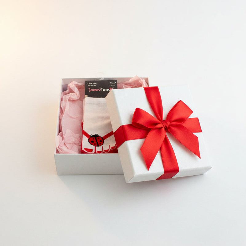 A pair of white baby socks with red trim and a red ladybug graphic on the sole, packaged on a black JazzyToes brand card against a white background.