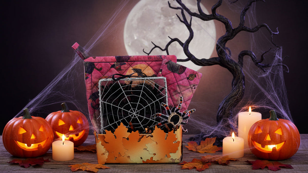 A Halloween-themed gift basket or box filled with seasonal items, including a pink patterned quilted pot holder, a black spiderweb square, a decorative spider, and an orange leaf-shaped base, photographed on a white background.