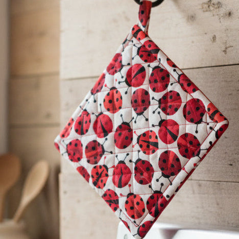 A quilted pot holder with a red and black ladybug pattern on a white background, hanging on a rustic wooden wall in a kitchen setting.