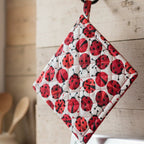 A quilted pot holder with a red and black ladybug pattern on a white background, hanging on a rustic wooden wall in a kitchen setting.