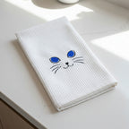 A folded white waffle-weave kitchen towel featuring a simple embroidered cat face with blue eyes, black whiskers, and a black nose on a plain white background.